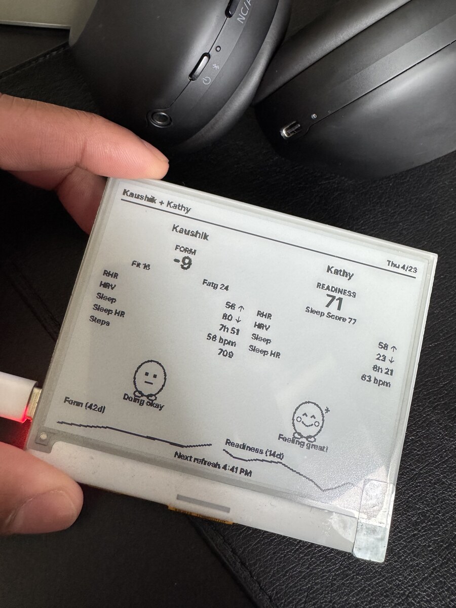 Handheld close-up of the Paperdink prototype showing the dual-athlete fitness page on its reflective e-ink panel.